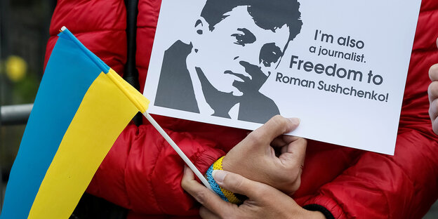 Eine Person hält eine ukrainische Flagge und ein Schild in der Hand mit der Aufschrift „I'm also a journalist. Freedom to Roman Sushchenko!““ Eine Person hält eine ukrainische Flagge und ein Schild in der Hand mit der Aufschrift „I'm also a journalist. Freedom to Roman Sushchenko!““