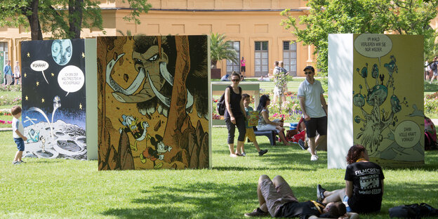 Besucher sitzen und liegen im Schloßpark. Um sie herum stehen große Wände mit Comics darauf. Besucher sitzen und liegen im Schloßpark. Um sie herum stehen große Wände mit Comics darauf.