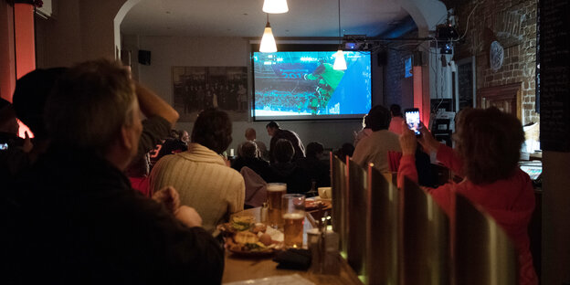 Menschen von hinten, die auf eine Leinwand mit Eishockey schauen, Pommes auf dem Tisch Menschen von hinten, die auf eine Leinwand mit Eishockey schauen, Pommes auf dem Tisch