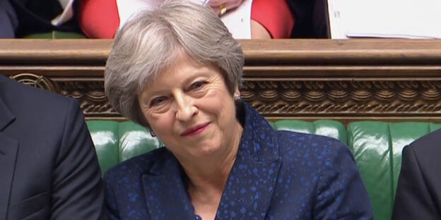 Theresa May, eine Frau mit kurzen grauen Haaren lächelt. Sie sitzt auf einer grün gepolsterten Bank. Eine Frau mit grauen kurzen Haaren spricht in ein Mikrofon, im Hintergrund gucken viele Menschen gespannt zu ihr Theresa May, eine Frau mit kurzen grauen Haaren lächelt. Sie sitzt auf einer grün gepolsterten Bank. Eine Frau mit grauen kurzen Haaren spricht in ein Mikrofon, im Hintergrund gucken viele Menschen gespannt zu ihr