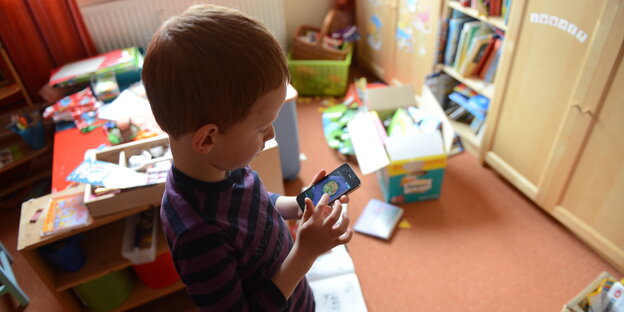 Ein Kind steht in einem Kinderzimmer mit Spielsachen und spielt auf seinem Smartphone Ein Kind steht in einem Kinderzimmer mit Spielsachen und spielt auf seinem Smartphone