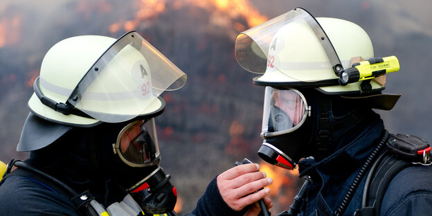 Zwei voll ausgerüstete Feuerwehrmänner blicken sich an Zwei voll ausgerüstete Feuerwehrmänner blicken sich an