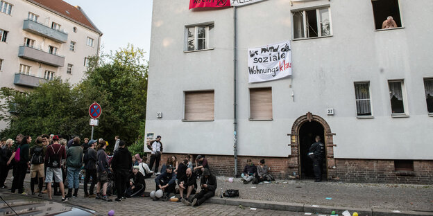 Jungen Leute sitzen vor einem Haus, in der Haustür steht ein Polizistavor, vor der Haustär