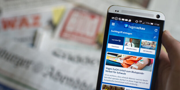 Auf einem Smartphone ist die Tagesschau-App geöffnet. Im Hintergrund liegen Zeitungen, wie beispielsweise die Bild Auf einem Smartphone ist die Tagesschau-App geöffnet. Im Hintergrund liegen Zeitungen, wie beispielsweise die Bild