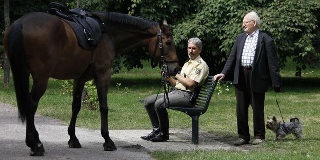 Ein Polizist sitzt auf einer Parkbank und hält ein Pferd am Halfter fest Ein Polizist sitzt auf einer Parkbank und hält ein Pferd am Halfter fest