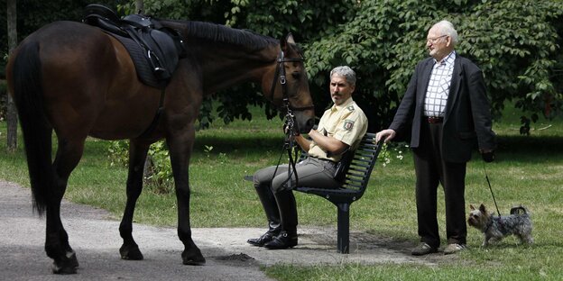 Ein Polizist sitzt auf einer Parkbank und hält ein Pferd am Halfter fest