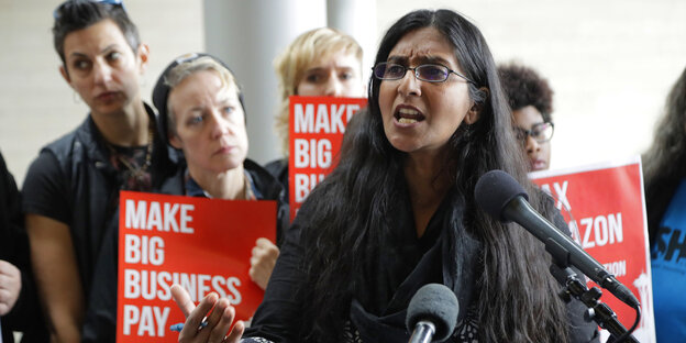 Seattles Stadträtin Kshama Sawant spricht. Im Hintergrund Menschen mit Schildern, auf denen steht: „Make Big Business Pay" Seattles Stadträtin Kshama Sawant spricht. Im Hintergrund Menschen mit Schildern, auf denen steht: „Make Big Business Pay"