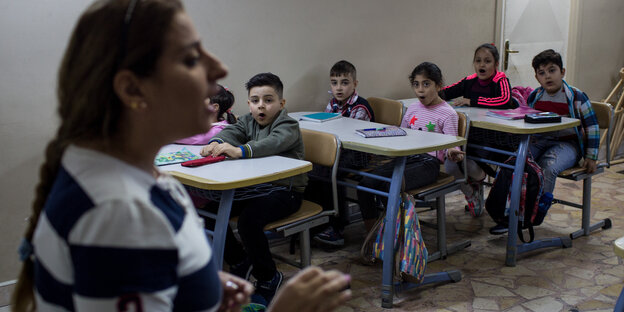 Kinder sitzen auf Schulbänken, vorne die Lehrerin Kinder sitzen auf Schulbänken, vorne die Lehrerin
