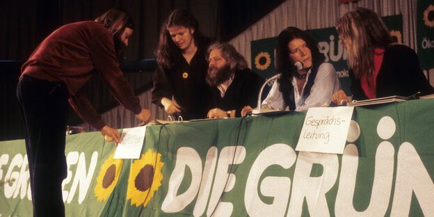 Verschiedene Menschen sitzen beim Parteitag der Grünen an einem Tisch, an dessen Vorderseite ein Transparent mit der Aufschrift „Die Grünen“ hängt Verschiedene Menschen sitzen beim Parteitag der Grünen an einem Tisch, an dessen Vorderseite ein Transparent mit der Aufschrift „Die Grünen“ hängt