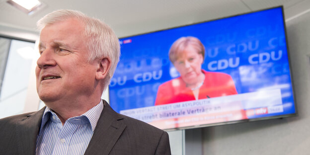 Horst Seehofer blickt nach links, hinter blickt Angela Merkel von einem Fernsehbildschirm herunter Horst Seehofer blickt nach links, hinter blickt Angela Merkel von einem Fernsehbildschirm herunter