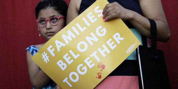 Ein Mädchen steht neben ihrer Mutter, die ein Schild mit der Aufschrift „Families belong together“ – „Familien gehören zusammen“ Ein Mädchen steht neben ihrer Mutter, die ein Schild mit der Aufschrift „Families belong together“ – „Familien gehören zusammen“