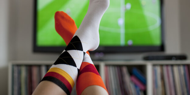 Ein Fernseher. Davor zu sehen: zwei Füße mit Socken in schwar-rot-gold Ein Fernseher. Davor zu sehen: zwei Füße mit Socken in schwar-rot-gold