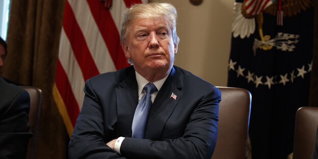 US-Präsident Trump sitzt im blauen Anzug mit verschränkten Armen vor einer US-Flagge US-Präsident Trump sitzt im blauen Anzug mit verschränkten Armen vor einer US-Flagge