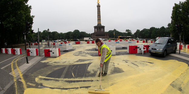 Ein Mann mit gelber Schutzweste trägt mit einem Besen gelbe Farbe auf die Straße auf, im HIntergrund die Siegessäule Ein Mann mit gelber Schutzweste trägt mit einem Besen gelbe Farbe auf die Straße auf, im HIntergrund die Siegessäule