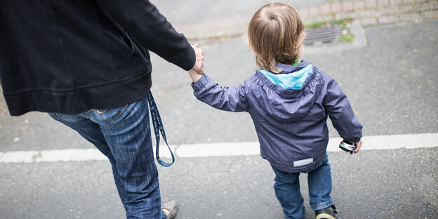 Eine Mutter hält ihr Kind an der Hand, beide sind von hinten zu sehen Eine Mutter hält ihr Kind an der Hand, beide sind von hinten zu sehen