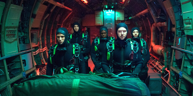 In einem U-Boot stehen fünf Navy-Seals in Taucheranzügen, oben leuchtet rotes Licht, vor den Personen liegt eine große Tasche In einem U-Boot stehen fünf Navy-Seals in Taucheranzügen, oben leuchtet rotes Licht, vor den Personen liegt eine große Tasche