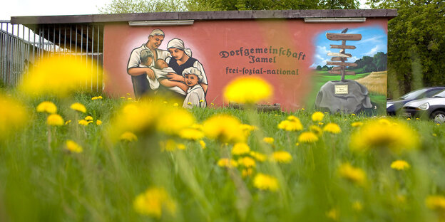 Auf einer Wiese blüht gelber Löwenzahn, dahinter steht auf einer braun-roten Scheunenwand in schwarzen, runenartigen Buchstaben: Dorfgemeinschaft Jamel - frei sozial national Auf einer Wiese blüht gelber Löwenzahn, dahinter steht auf einer braun-roten Scheunenwand in schwarzen, runenartigen Buchstaben: Dorfgemeinschaft Jamel - frei sozial national