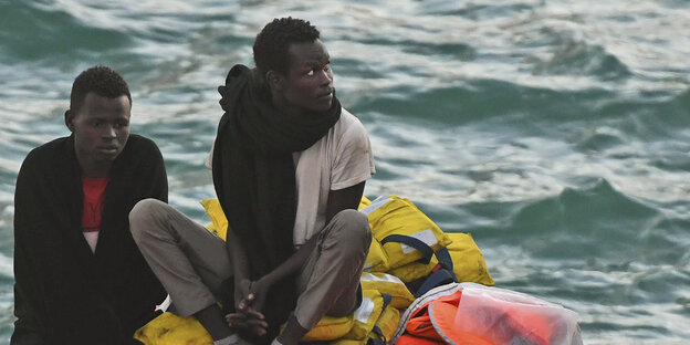 Zwei Geflüchtete sitzen auf Rettungswesten auf einem Boot. Zwei Geflüchtete sitzen auf Rettungswesten auf einem Boot.