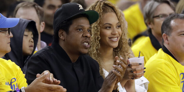 Jay-Z und Beyoncé sitzen nebeneinander in einem Basketball-Stadion und trinken Bier Jay-Z und Beyoncé sitzen nebeneinander in einem Basketball-Stadion und trinken Bier