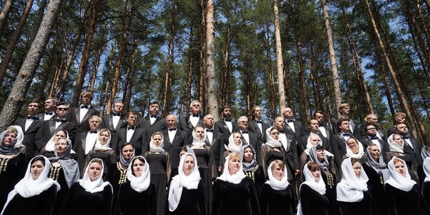 Ein Chor steht vor hohen Kiefern, alle sind schwarz gekleidet und haben das Haupt mit weißen Tüchern bedeckt Ein Chor steht vor hohen Kiefern, alle sind schwarz gekleidet und haben das Haupt mit weißen Tüchern bedeckt