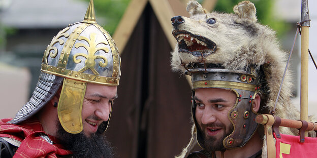 Zwei Männerköpfe mit Helmen, einer davon trägt auf seinem Helm auch noch einen ausgestopften Bärenkopf Zwei Männerköpfe mit Helmen, einer davon trägt auf seinem Helm auch noch einen ausgestopften Bärenkopf