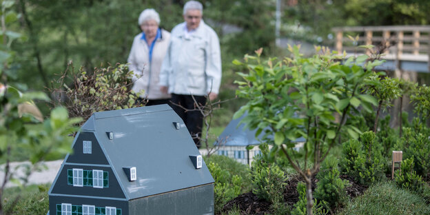 Im Vordergrund steht ein Miniaturhaus, im Hintergrund läuft ein älteres Paar Im Vordergrund steht ein Miniaturhaus, im Hintergrund läuft ein älteres Paar