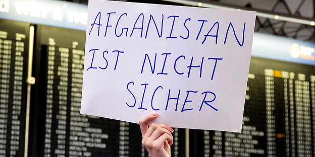 Vor einer Anzeigetafel am Flughafen hält eine Person ein Schild „Afghanistan ist nicht sicher“ Vor einer Anzeigetafel am Flughafen hält eine Person ein Schild „Afghanistan ist nicht sicher“