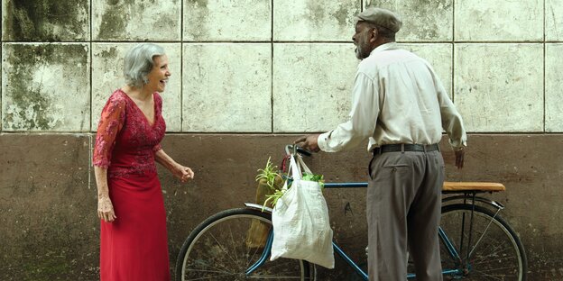 Das foto zeigt die beiden Hauptfiguren Candelaria und Victor vor einer grauen Hauswand. Die beiden lachen sich an. Er hält ein Fahrrad in der Hand, sie trägt ein rotes Kleid