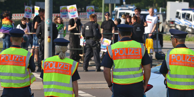 Polizist*innen stehen mit neongelben Warnwesten bekleidet an einer Absperrung. Dahinter sieht man Demonstranten Polizist*innen stehen mit neongelben Warnwesten bekleidet an einer Absperrung. Dahinter sieht man Demonstranten