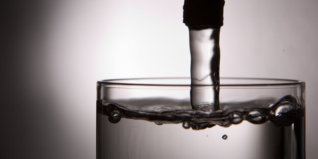 Aus einem Wasserhahn fließt Wasser in ein schon fast bis obenhin gefülltes Glas Aus einem Wasserhahn fließt Wasser in ein schon fast bis obenhin gefülltes Glas