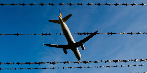 Blick durch Stacheldraht auf ein am Himmel fliegendes Flugzeug