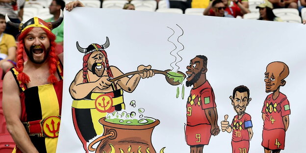 ein als Obelix in den belgischen Nationalfarben verkleideter Mann hält ein Transparent, auf dem Obelix Zaubertrank an belgische Nationalspieler ausschenkt ein als Obelix in den belgischen Nationalfarben verkleideter Mann hält ein Transparent, auf dem Obelix Zaubertrank an belgische Nationalspieler ausschenkt