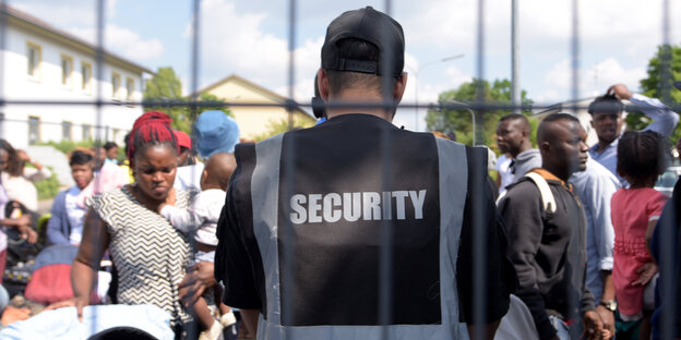 Ein Mann mit einer Security-Weste steht mit dem Rücken an ein Gitter gelehnt, sein Gesicht ist nicht zu sehen, wohl aber die Gesichter wartender Flüchtlinge, die vor ihm stehen Ein Mann mit einer Security-Weste steht mit dem Rücken an ein Gitter gelehnt, sein Gesicht ist nicht zu sehen, wohl aber die Gesichter wartender Flüchtlinge, die vor ihm stehen