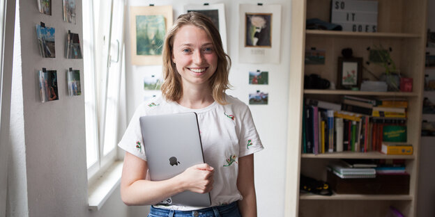 Eine lächelnde junge Frau mit Laptop unter Arm steht neben einem Bücherregal