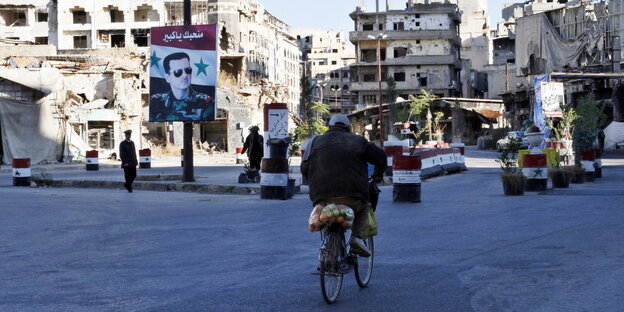 Im syrischen Homs fährt ein Mann auf dem Fahrrad auf einen Kontrollposten zu. Im Hintergrund ausgebombte Häuser und ein Bild des Präsidenten Assad Im syrischen Homs fährt ein Mann auf dem Fahrrad auf einen Kontrollposten zu. Im Hintergrund ausgebombte Häuser und ein Bild des Präsidenten Assad