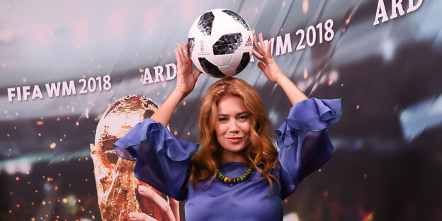 eine Frau hält einen Fußball über den Kopf, im Hintergrund eine Studiowand mit einer Abbildung des WM-Pokals und dem Schriftzug „Fifa WM 2018 ARD“ eine Frau hält einen Fußball über den Kopf, im Hintergrund eine Studiowand mit einer Abbildung des WM-Pokals und dem Schriftzug „Fifa WM 2018 ARD“