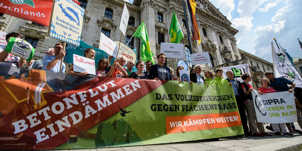 Demonstranten halten Transparente hoch, auf denen steht: Betonflut eindämmen. Das Volksbegehren gegen Flächenfraß Demonstranten halten Transparente hoch, auf denen steht: Betonflut eindämmen. Das Volksbegehren gegen Flächenfraß