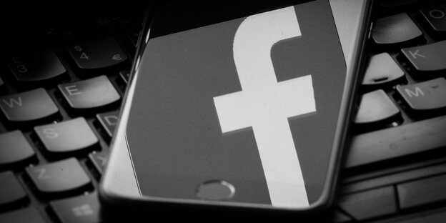 Ein Handy, in dem sich das Facebook-Logo spiegelt, liegt auf einer Tastatur, alles ist schwarz-weiß Ein Handy, in dem sich das Facebook-Logo spiegelt, liegt auf einer Tastatur, alles ist schwarz-weiß
