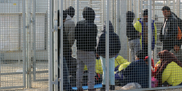Eine Gruppe von Menschen in Jacken steht und sitzt in einem eingezäunten Bereich Eine Gruppe von Menschen in Jacken steht und sitzt in einem eingezäunten Bereich
