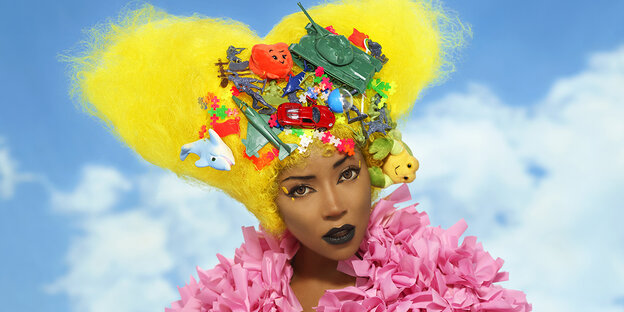 Ebony Bones trägt eine blonde Perücke, in der sich verschiedene Spielzeuge befinden Ebony Bones trägt eine blonde Perücke, in der sich verschiedene Spielzeuge befinden