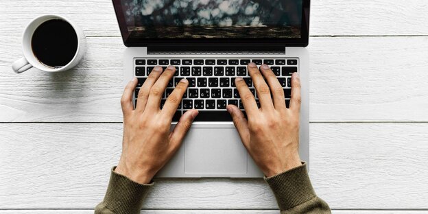 ZWei Hände tippten auf einer Laptop-Tastatur. Daneben steht eine Tassee Kaffee ZWei Hände tippten auf einer Laptop-Tastatur. Daneben steht eine Tassee Kaffee