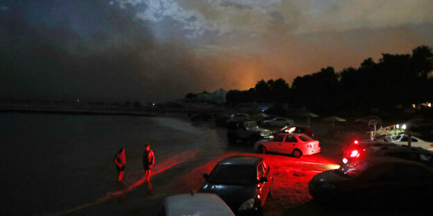 Autos stehen an einem Strand, es ist Nacht, im Hintergrund brennen Wälder