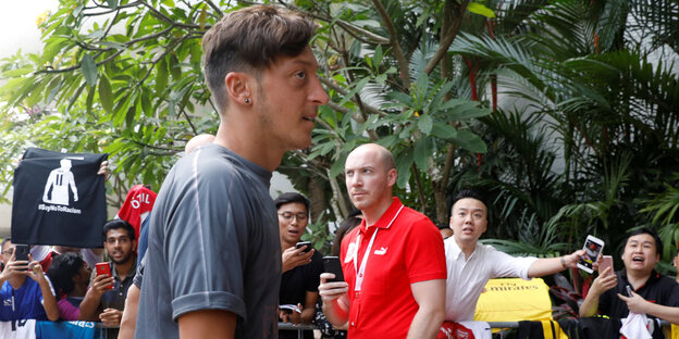 Menschen in roten T-Shirts stehen im Hintergrund, Mesut Özil trägt ein graues Shirt und wendet sich ab Menschen in roten T-Shirts stehen im Hintergrund, Mesut Özil trägt ein graues Shirt und wendet sich ab