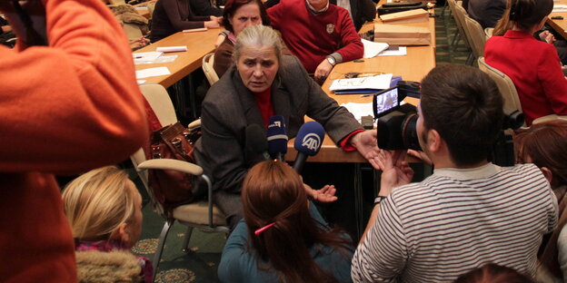 Eine Frau sitzt inmitten eines vollen Konferenzraums und spricht in Mikrofone Eine Frau sitzt inmitten eines vollen Konferenzraums und spricht in Mikrofone