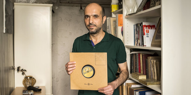 Ein Mann steht zwischen Regalen in seiner Wohnung und hält eine Schallplatte in altem braunen Papier in den Händen Ein Mann steht zwischen Regalen in seiner Wohnung und hält eine Schallplatte in altem braunen Papier in den Händen