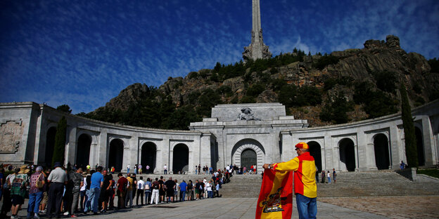 Vor einem Mausoleum stehen Personen an, daneben steht eine Person mit der Flagge Spaniens aus der Endphase der Diktatur Francos Vor einem Mausoleum stehen Personen an, daneben steht eine Person mit der Flagge Spaniens aus der Endphase der Diktatur Francos