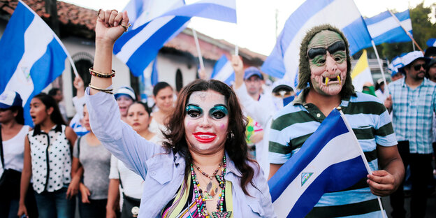 Eine übertrieben stark geschminkte Frau und ein mit einer Horrorfratze bemalter Mann marschieren bei einer Demonstration mit nicaraguanischen Fahrnen Eine übertrieben stark geschminkte Frau und ein mit einer Horrorfratze bemalter Mann marschieren bei einer Demonstration mit nicaraguanischen Fahrnen