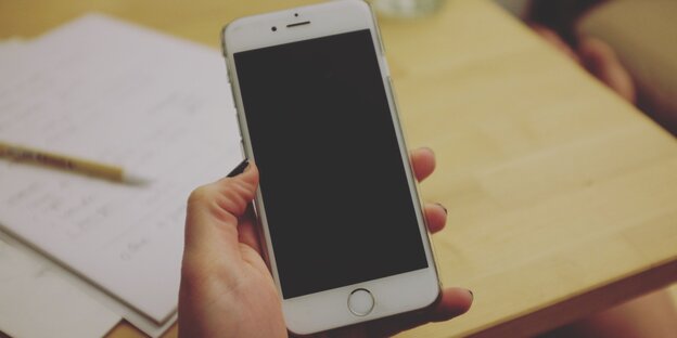 JEmand hält ein Handy in der HAnd. Dahinter sieht man einen Tisch, auf dem Zettel liegen JEmand hält ein Handy in der HAnd. Dahinter sieht man einen Tisch, auf dem Zettel liegen