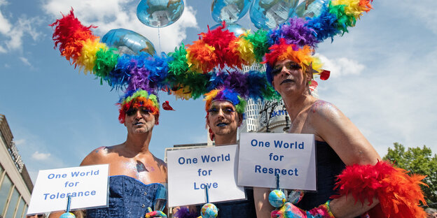 Drei Personen mit Regenbogenpuscheln auf dem Haar und Schildern, die weltweite Toleranz fordern Drei Personen mit Regenbogenpuscheln auf dem Haar und Schildern, die weltweite Toleranz fordern
