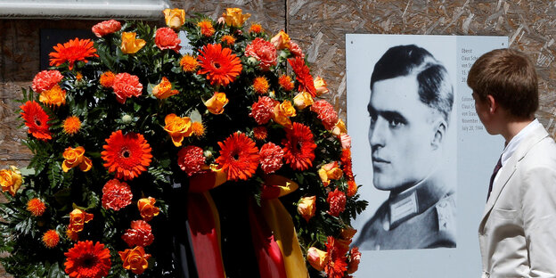 Eine Person steht vor einem Porträt von Graf Stauffenberg, sie ist nur von hinten zu sehen, neben dem Porträt sind Blumen drapiert Eine Person steht vor einem Porträt von Graf Stauffenberg, sie ist nur von hinten zu sehen, neben dem Porträt sind Blumen drapiert